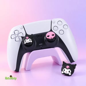 Puede incluir: Un mando de videojuegos blanco y negro sobre un soporte de color violeta claro. El mando incluye dos fundas para los joysticks: una con una cara de personaje negra y una calavera rosa, y otra con forma de calavera rosa. Una funda a juego está al lado.