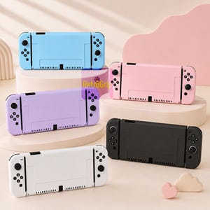 May include: Five handheld gaming consoles in light blue, pastel pink, lavender, white, and black. Each device features a central screen area with detachable controllers on both sides, including directional pads, action buttons, and analog sticks.