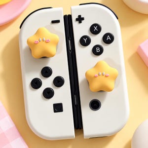 Puede incluir: Un par de mandos de juego blancos con botones negros y dos fundas para joystick en forma de estrella amarilla. Cada funda de estrella tiene una cara pequeña y sencilla con mejillas rosas y ojos blancos, unida a los sticks analógicos.