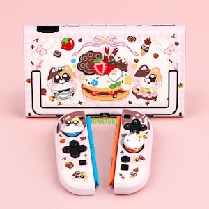 May include: A light pink gaming console and two controllers featuring a dessert-themed pattern with cartoon cats, strawberries, chocolate, and a large central cake illustration. The text RetroGrip is printed in green between the two controllers.