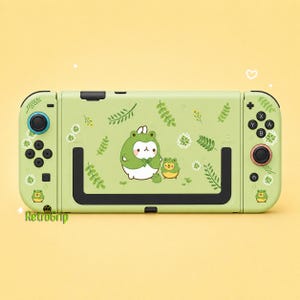 May include: A light green handheld gaming console featuring a central graphic of a white character in a frog costume alongside a smaller yellow frog character. The console is decorated with small green leaf and white flower patterns and the text RetroGrip.