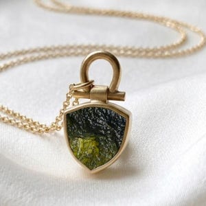 May include: A gold necklace featuring a shield-shaped pendant with a textured, dark green, translucent stone set in a gold frame. The pendant is attached to a delicate gold chain by a U-shaped gold bail, creating a cohesive and refined jewellery piece.