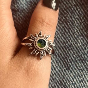 Könnte beinhalten: Ein silberfarbener Ring mit einem Sonnenstrahlen-Design und einem runden grünen Stein in der Mitte. Die Ringschiene ist zweigeteilt und die Sonnenstrahlen sind entlang der Kanten des Metallrahmens mit kleinen Punkten verziert.