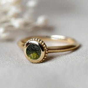 Op de afbeelding: Een gouden ring met een ronde zetting met een textuurrand en een donkergroene, ruwe steen in het midden. De ringband is glad en dun. De tekst AnviJewelsStudio is in kleine zwarte letters zichtbaar aan de zijkant van de zetting van de ring.