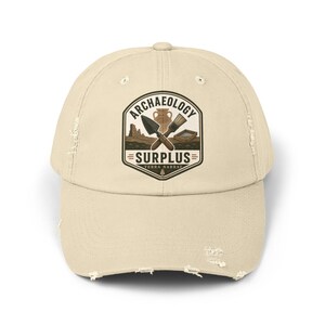 May include: A beige distressed baseball cap featuring a central graphic emblem with the text ARCHAEOLOGY SURPLUS and TERRA NARRAT. The design includes illustrations of a trowel, a brush, a pottery vessel, and a sifting screen on a dark ground.