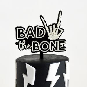 Pode incluir: Um topo de bolo em preto e branco com o texto BAD to the BONE em fonte negrito, acompanhado por uma mão de esqueleto branca fazendo um gesto de rock and roll. O topo está posicionado sobre um bolo preto decorado com raios brancos.