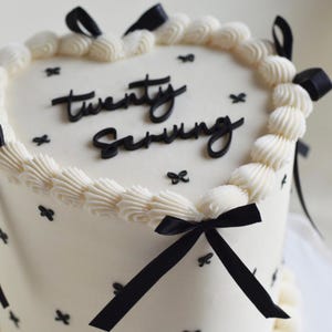 Op de afbeelding: Een hartvormige taart met glad wit glazuur, versierd met zwarte strikjes en zwarte lintaccenten. De bovenkant bevat de zwarte tekst "twenty serving" in schrijfletters en een rand van witte toeven glazuur langs de zijkant.