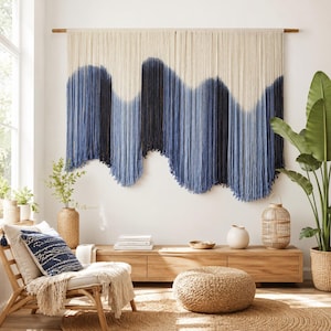 May include: A large wall hanging featuring long, vertical cream-colored cotton cords suspended from a wooden rod. The lower sections of the cords are dyed in a gradient of blue tones, creating a wavy, mountain-like pattern across the width.