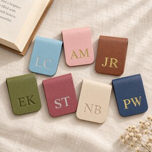 May include: A collection of seven rectangular magnetic bookmarks made of faux leather in various colors including light blue, pink, brown, olive green, maroon, cream, and navy blue, each featuring two embossed or foil-stamped capital letters.