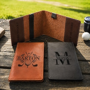 May include: Two rectangular leather golf scorecards, one tan and one black, featuring custom engravings. The tan cover displays the name BARTON with golf clubs and a ball. The black cover shows the name Marvin with a large letter M. The interior is brown.