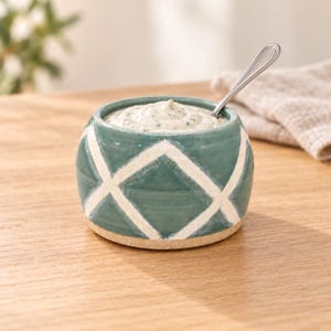 May include: A round, teal ceramic bowl featuring a white crisscross diamond pattern. The bowl is filled with a creamy, speckled white dip and includes a small, polished silver metal spoon resting inside the container.