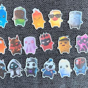 May include: A collection of seventeen small, colorful stickers featuring various cartoon characters with distinct designs, including creatures with horns, masks, sunglasses, and elemental themes, arranged in three rows on a dark gray textured surface.