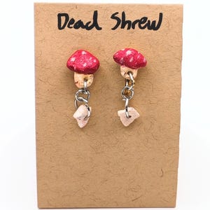 May include: A pair of earrings featuring red mushroom caps with white spots and tan stems, attached to silver-toned metal rings with dangling light-coloured stone fragments. The earrings are displayed on a tan card with the text Dead Shrew written in black.