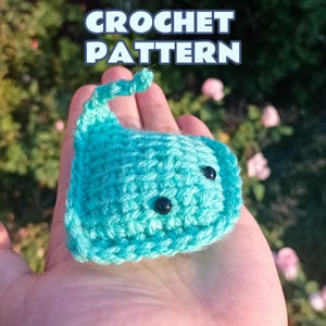 May include: A small, turquoise crochet stingray plushie featuring two black bead eyes and a curved tail. The text CROCHET PATTERN is displayed in white letters with a dark blue outline at the top of the image, indicating a digital design guide.