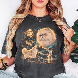 May include: A charcoal gray short-sleeve t-shirt featuring a graphic print of a fluffy cat, a full moon, a forest, and a waterfall. The design includes a large cat face in the sky above a smaller standing cat in a landscape setting.