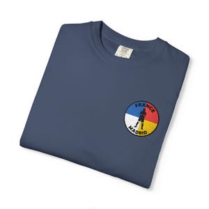 May include: A navy blue t-shirt featuring a circular embroidered patch on the chest. The patch is divided into four colored quadrants of blue, red, yellow, and white, with the text "FRANCE" and "MADRID" and a silhouette of a soccer player.