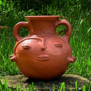 Puede incluir: Una vasija de cerámica color terracota con un cuerpo redondeado y un rostro humano esculpido, que incluye ojos cerrados, nariz y labios. La pieza tiene dos asas simétricas que se extienden desde el cuello hacia los lados del cuerpo.