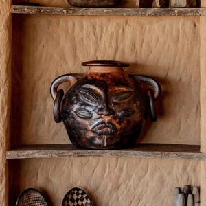 May include: A dark brown ceramic vessel with two handles and a sculpted face featuring multiple eyes and lips, displayed on a shelf alongside two small patterned bowls and a collection of grey stone-like objects in a textured earthen wall niche.