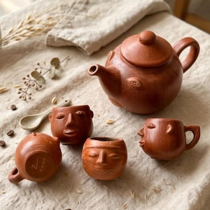 May include: A terracotta tea set featuring a round teapot with a lid and three small cups, each sculpted with human facial features. The teapot body displays embossed eye motifs. One cup shows the text "Zu o'o.co" on its base. A small spoon is included.