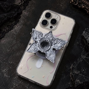 Puede incluir: Una funda transparente para smartphone con un accesorio plateado en forma de flor de varios pétalos, con una abertura circular central bordeada por pequeñas protuberancias afiladas similares a dientes, situada en la parte trasera del dispositivo.