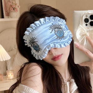 Puede incluir: Un antifaz de dormir de satén azul claro con un borde con volantes y dos diseños en forma de ojo adornados con pedrería negra y azul. El antifaz está confeccionado con una tela suave y brillante e incluye un ribete fruncido en todo el borde.