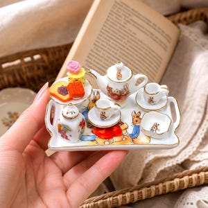 May include: A miniature white ceramic tea set on a rectangular tray featuring Peter Rabbit illustrations. The set includes a teapot, two teacups with saucers, a lidded sugar bowl, a small square dish, and a plate with tiny food items like a cake.