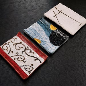 May include: Three rectangular ceramic trays with distinct designs. One features a white surface with black line art, another has a blue and black textured pattern with yellow accents, and the third has a white center with black swirls and red borders.