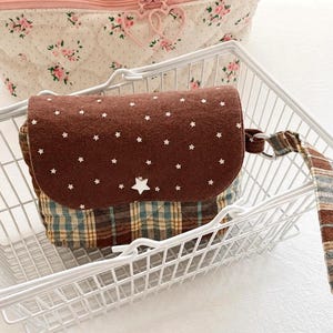 May include: A small rectangular pouch with a brown flap featuring white star patterns and a single white star button. The body of the pouch is made of quilted fabric with a blue, brown, and yellow plaid pattern. It includes a matching plaid wrist strap.