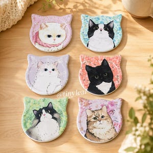 May include: A set of six ceramic cat-shaped coasters, each featuring a unique illustration of a cat against a floral background. The designs include white, black, and tabby cats in various colours, with the text @tinyleaf visible in the centre.