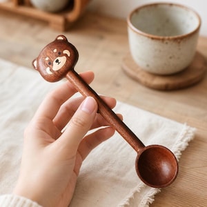 Può includere: Un cucchiaio in legno marrone scuro con una testa di panda rosso intagliata sulla parte superiore del manico. Il cucchiaio presenta un manico lungo e sottile e una piccola coppa circolare all'estremità. La venatura del legno è visibile.