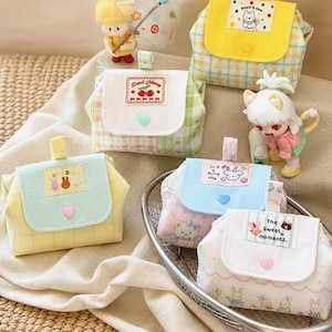 Pode incluir: Cinco pequenas bolsas de tecido com fechos de aba, cada uma com padrões xadrez pastel, botões em forma de coração e etiquetas retangulares com ilustrações e textos como "Sweet Cherry", "It's a Bunny day" e "The Sweet moments".
