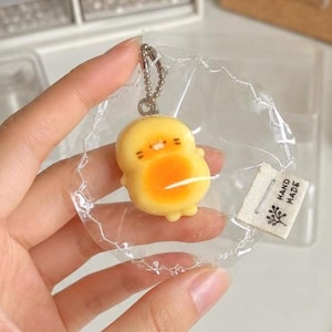 Peut inclure: Une petite breloque personnage jaune avec un corps arrondi, deux yeux et une bouche, attachée à une chaînette à billes argentée. La breloque est enfermée dans un sachet plastique transparent avec une étiquette en tissu blanc marquée HAND MADE.