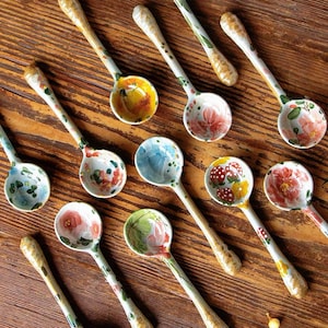 May include: A collection of eleven ceramic spoons featuring hand-painted floral, fruit, and botanical motifs in various colours including pink, red, yellow, and green. Each spoon has a long, slender handle and a rounded bowl with unique designs.