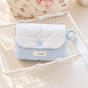 Puede incluir: Un bolso acolchado de color azul claro con una solapa blanca con estampado de puntos azules. El bolso tiene un cierre de botón azul en forma de corazón, una correa de muñeca a juego y una etiqueta de tela con el texto hand made.
