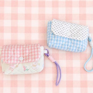 May include: Two quilted fabric pouches with snap closures and wrist straps. One pouch features a pink gingham flap over a white base with small floral prints. The other pouch has a blue gingham base with a white flap featuring black polka dots.