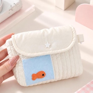 Puede incluir: Un pequeño estuche rectangular acolchado de color crema con cierre de solapa y un botón blanco en forma de estrella. En la parte frontal lleva cosido un cuadrado azul claro con una aplicación de pez naranja y un detalle de ojo negro.