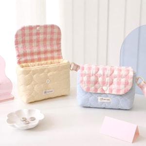 May include: Two small quilted crossbody bags with heart-shaped stitching. One bag is cream with a pink and white gingham flap, and the other is light blue with a matching pink and white gingham flap. Both feature a small white label with the text Hand Made.