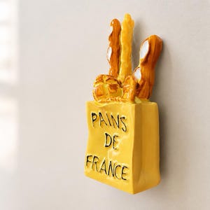 Puede incluir: Un soporte de pared de cer&aacute;mica amarilla con forma de bolsa de papel llena de pan. La bolsa presenta el texto en relieve negro PAINS DE FRANCE. Los panes est&aacute;n representados en tonos marr&oacute;n dorado y amarillo con un acabado brillante y liso.
