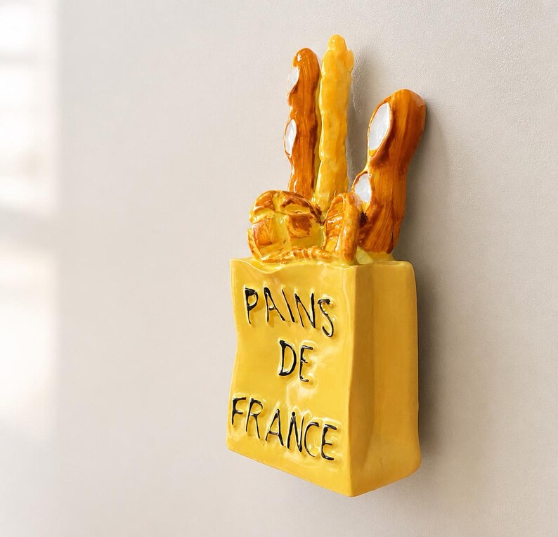 Puede incluir: Un soporte de pared de cer&aacute;mica amarilla con forma de bolsa de papel llena de pan. La bolsa presenta el texto en relieve negro PAINS DE FRANCE. Los panes est&aacute;n representados en tonos marr&oacute;n dorado y amarillo con un acabado brillante y liso.