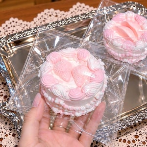 Puede incluir: Una pequeña vela redonda con forma de pastel decorada con remolinos de glaseado rosa y blanco y dos detalles en forma de fresa rosa en la parte superior. La vela está envuelta en un plástico cuadrado transparente y tiene una base texturizada.