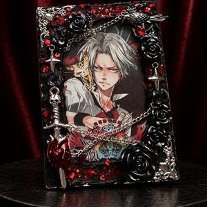 Puede incluir: Un marco de fotos rectangular con una ilustración anime central de un personaje con cabello plateado puntiagudo. El marco está decorado con rosas negras, dijes de metal plateado que incluyen una espada, una calavera y estrellas, y detalles de purpurina roja.