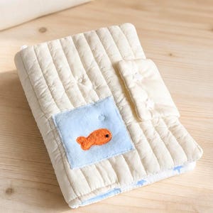 May include: A cream-colored quilted fabric book cover featuring vertical stitching and a light blue square patch on the front. The patch displays an orange fish appliqué. A matching quilted flap with star-shaped stitching secures the cover.