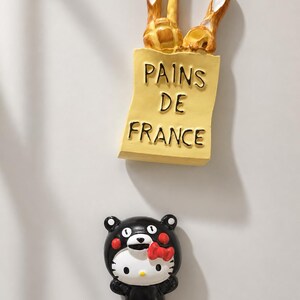 Puede incluir: Un im&aacute;n de nevera con forma de bolsa de papel amarilla con la etiqueta PAINS DE FRANCE que contiene tres barras de pan doradas, y un segundo im&aacute;n de nevera con un personaje de gato blanco con un traje de oso negro y un lazo rojo.