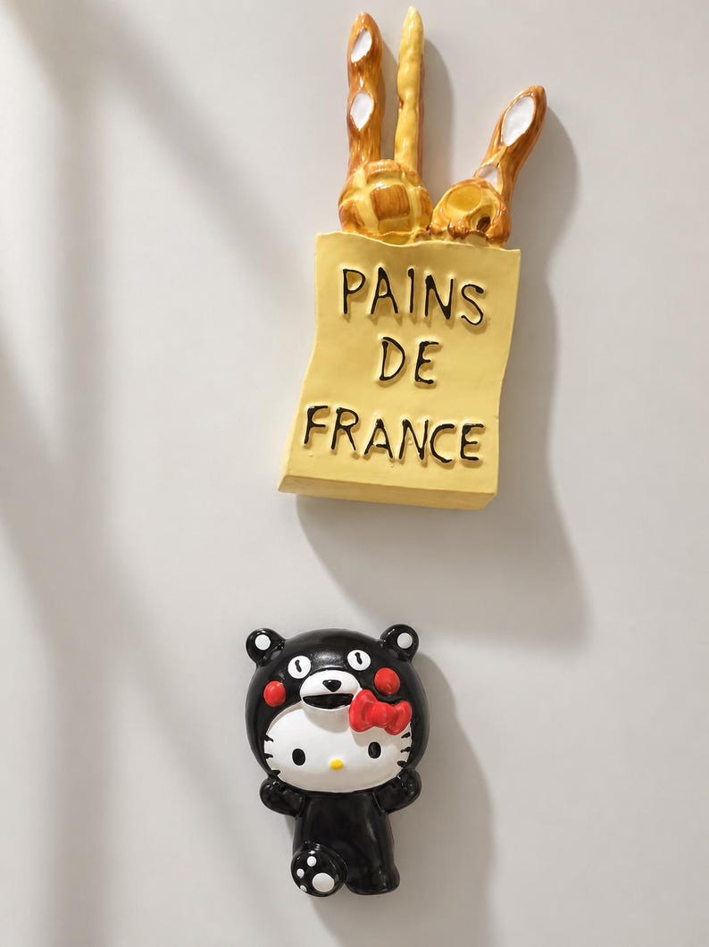 Puede incluir: Un im&aacute;n de nevera con forma de bolsa de papel amarilla con la etiqueta PAINS DE FRANCE que contiene tres barras de pan doradas, y un segundo im&aacute;n de nevera con un personaje de gato blanco con un traje de oso negro y un lazo rojo.