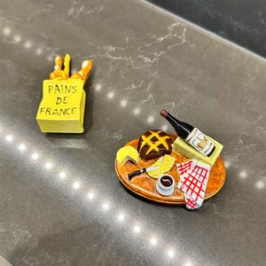 Puede incluir: Dos art&iacute;culos decorativos en miniatura: una bolsa de papel amarilla con la etiqueta PAINS DE FRANCE que contiene pan, y una tabla de madera ovalada con una hogaza oscura, un trozo de queso amarillo, un cuchillo, una botella de vino tinto y una servilleta.