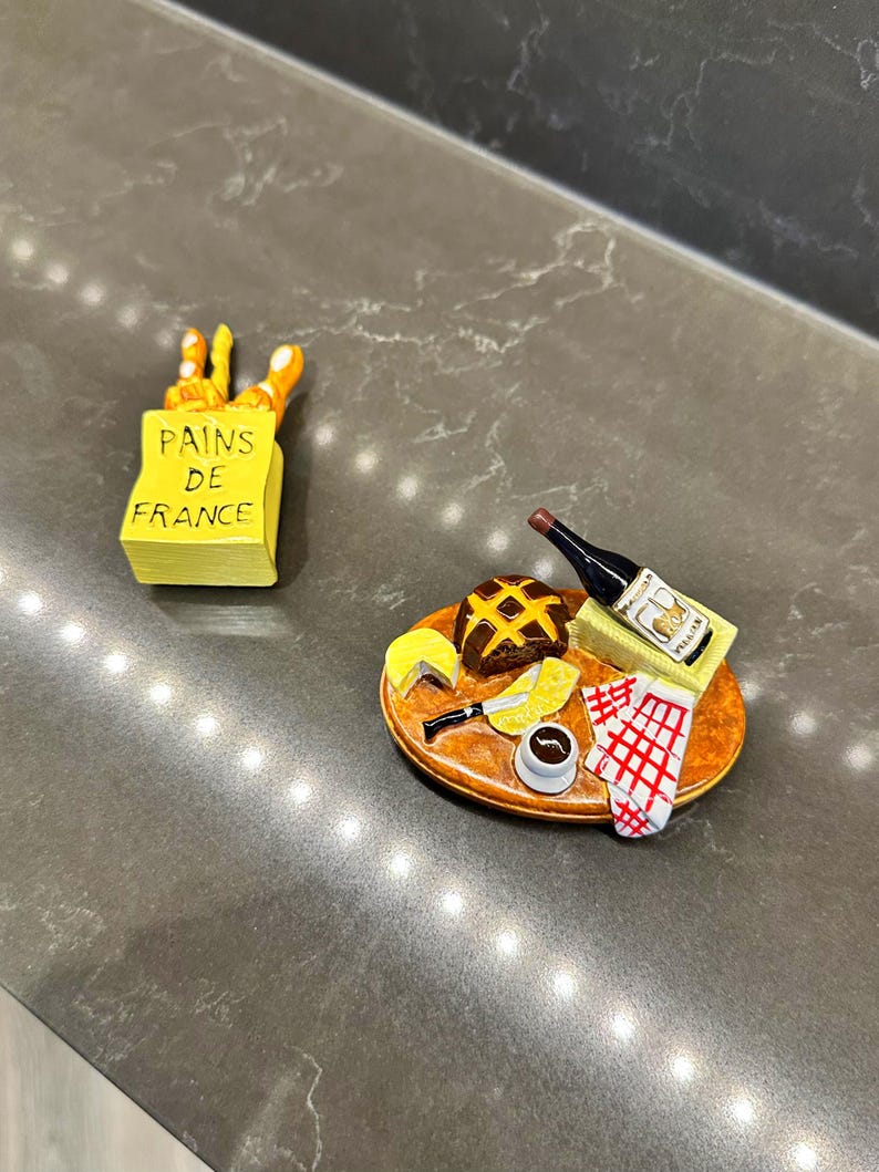 Puede incluir: Dos art&iacute;culos decorativos en miniatura: una bolsa de papel amarilla con la etiqueta PAINS DE FRANCE que contiene pan, y una tabla de madera ovalada con una hogaza oscura, un trozo de queso amarillo, un cuchillo, una botella de vino tinto y una servilleta.