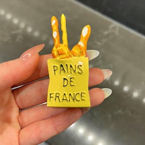 Puede incluir: Una peque&ntilde;a figura rectangular amarilla con forma de bolsa de papel que contiene tres baguettes de color marr&oacute;n dorado. El frente de la bolsa presenta el texto negro PAINS DE FRANCE impreso en una fuente clara y sin remates. Acabado brillante.