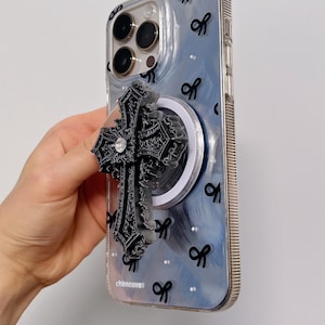 Puede incluir: Una funda transparente para smartphone con fondo marmolado azul y blanco, pequeños iconos de lazos negros y el texto chancuxe. En la parte posterior hay un soporte para teléfono negro, ornamentado y en forma de cruz con un brillante.