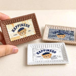May include: Three rectangular trays with pleated edges in brown, beige, and white. Each tray features a central graphic of a bear wearing a blue beret with the text "HAPPINESS SINCE 1988" printed in dark blue letters over a newspaper-style background.
