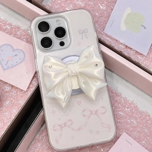 Può includere: Una custodia trasparente per smartphone con un grande fiocco tridimensionale in raso color crema con piccoli cristalli incastonati. La superficie della custodia è decorata con una delicata stampa grafica rosa chiaro di nastri, cuori e stelle.